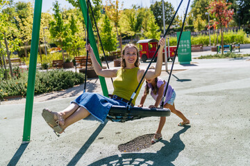 Mother and daughter smiling, playing on a swing together in a sunny urban park, enjoying outdoor family fun