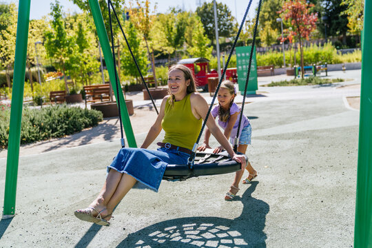 Happy mother and daughter enjoying a sunny day, laughing and playing on a swing set at an urban playground - Powered by Adobe