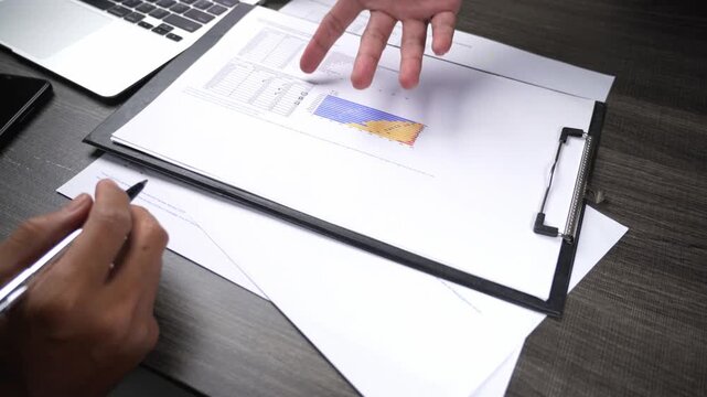 Hand making a gestural movement over a data document filled with charts and graphs, seen on a desk with a laptop and smartphone