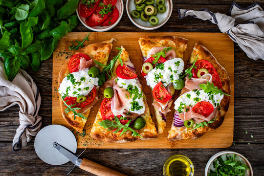 Pinsa with cured ham, burrata cheese, tomatoes, green olives and arugula on wooden table. Top view	
