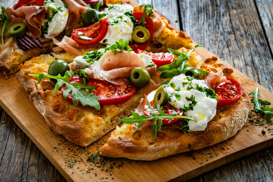Pinsa with cured ham, burrata cheese, tomatoes, green olives and arugula on wooden table	
