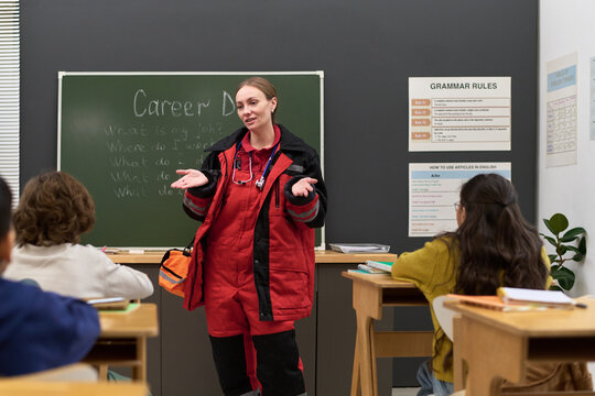 Caucasian young adult woman paramedic standing in front of classroom talking to group of children sitting at desks, blackboard with career day topic and grammar rules poster visible