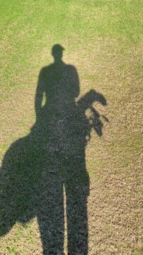 A person is standing in the grass with a golf club and a golf bag. The shadow of the person is cast on the ground