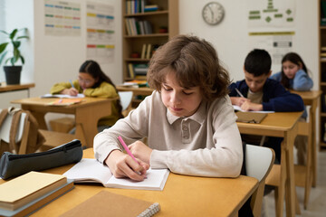 Caucasian boy teenager sitting at desk writing in notebook in classroom with multiethnic classmates studying in background, focused on schoolwork during lesson