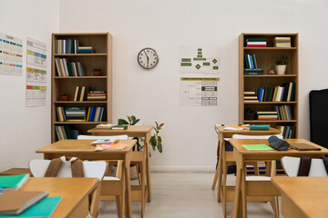 Empty classroom showing wooden desks and chairs arranged in rows, bookshelves filled with books, wall clock, educational posters on walls, potted plants adding decor, no people present