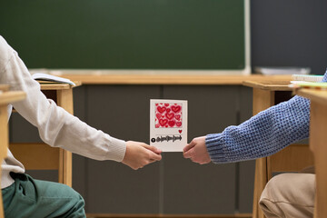 Two teenagers sitting at desks exchanging handmade card decorated with red hearts and soundwave drawing in classroom, only hands and arms of Caucasian and Black teenagers visible
