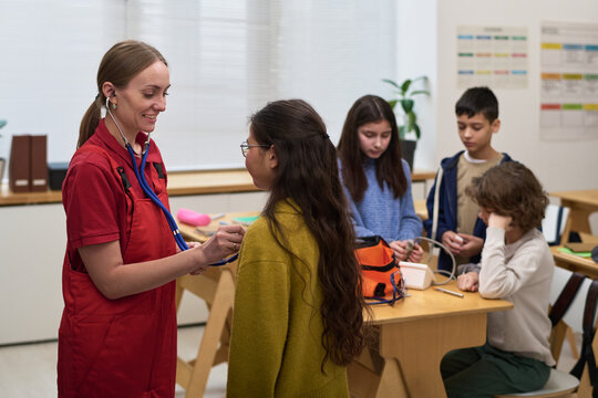 Caucasian young adult woman using stethoscope examining Asian preteen girl, while diverse group of children sitting at table engaging with medical equipment in classroom setting