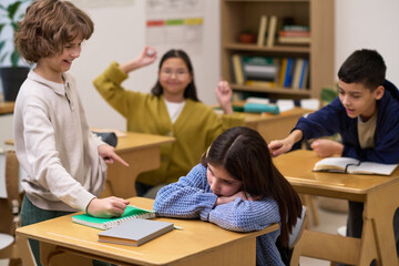 Caucasian girl sitting at desk with head down being bullied by group of classmates including Caucasian boy, Asian girl, and Caucasian boy pointing and laughing in classroom