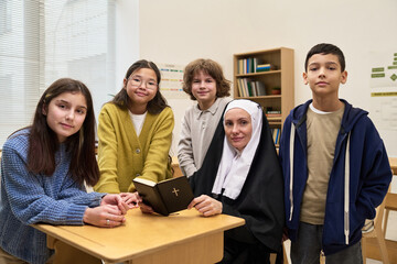 Caucasian nun sitting at desk holding Bible surrounded by diverse group of children and teenagers standing in classroom setting, all looking at camera with neutral expressions