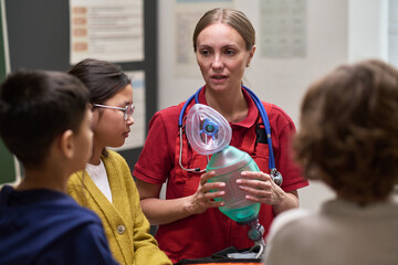 Caucasian young adult woman demonstrating medical resuscitator mask to group of attentive children in classroom setting, children watching closely as instructor explains equipment