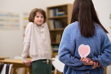 Teenage girl with brown hair standing with back to camera holding paper heart behind back, while teenage Caucasian boy with curly hair sitting at desk looking at her in classroom