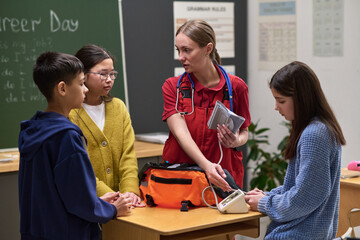 Caucasian young adult woman nurse demonstrating blood pressure measurement to diverse group of three teenagers in classroom setting, using medical equipment on wooden desk