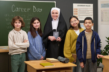 Caucasian nun standing in classroom holding Bible surrounded by diverse group of four children smiling, blackboard with Career Day written in background, desks and educational materials visible