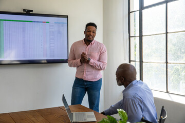 African American male coworkers presenting spreadsheet on wall monitor in compact office room