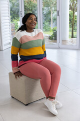 Naklejka premium African American woman sitting on beige ottoman by glass doors, wearing striped knit, smiling up