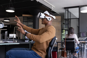 Worker wearing vr headset gesturing by lamp as coworker typing on laptop in open plan office