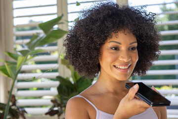 African american woman in tank top holding smartphone at home next to shutters by potted plants