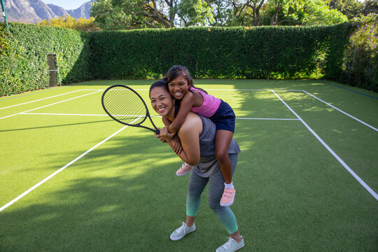 Asian mother and daughter giving piggyback ride on green tennis court with white lines holding rac