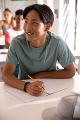 Diverse teenage classmates sitting at desks in classroom, writing in notebooks with pencil © wavebreak3