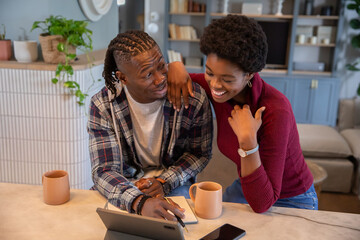 African american couple writing in notebook and leaning at home countertop table with mugs