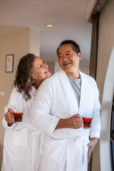 Diverse couple wearing bathrobes and holding amber tea mugs in softly lit living area