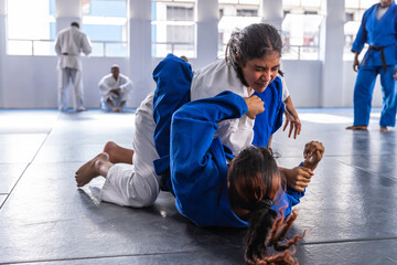 Asian teenage girls grappling on padded mats under windows inside dojo wearing white, blue gis