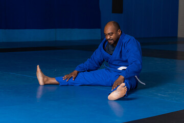 Senior african american man performing straddle stretch in gi on mats in dojo