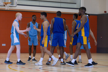 Diverse male basketball players shaking hands in gymnasium under basketball hoop