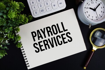 Payroll Services text written on notepad with alarm clock, magnifying glass and calculator on black background