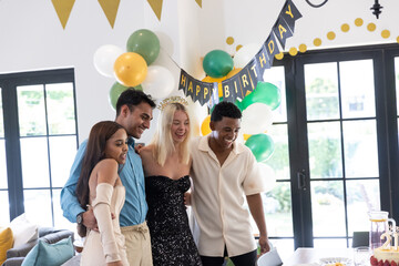 Diverse friends standing at home dining table under happy birthday banner with balloons, cake