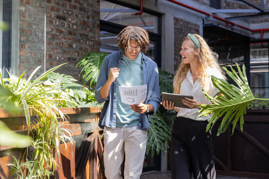 Diverse coworkers walking office hallway reviewing printed report and tablet with glasses by plants - Powered by Adobe