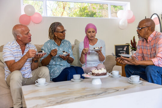 Diverse friends celebrating in living room with cake on coffee table, cancer survivor sign