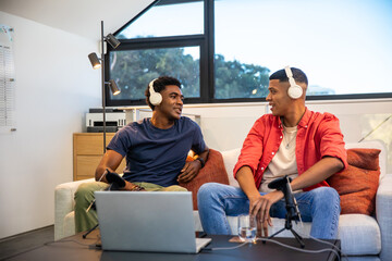 Diverse male friends recording podcast chatting on loveseat in home studio with laptop mic
