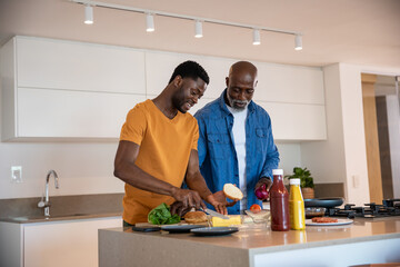 African american father and son slicing buns arranging cheese lettuce on island, pan, in kitchen