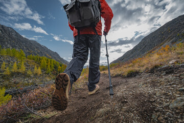 Low angle view of a person hiking up a rocky mountain trail during autumn, wearing a red jacket, black trousers, and using a trekking pole. © ANDREY