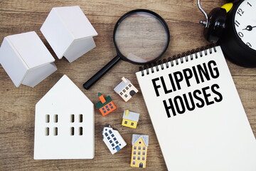 Flipping Houses text written on notepad with house model, magnifying glass and alarm clock on wooden background