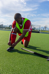 Obraz premium African american man kneeling and tying cleat on turf field beside fluorescent bib, hockey stick