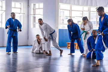 Diverse martial arts class practicing grappling drills on padded mats in dojo with gis, belts