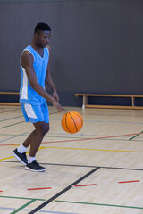 Naklejka premium African american man in basketball jersey dribbling ball on court by bench in gym, copy space