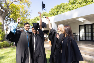 Diverse classmates in academic gowns tossing mortarboard caps celebrating graduation on lawn