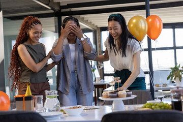 Diverse coworkers guiding surprised colleague toward cupcake stand at modern office celebration