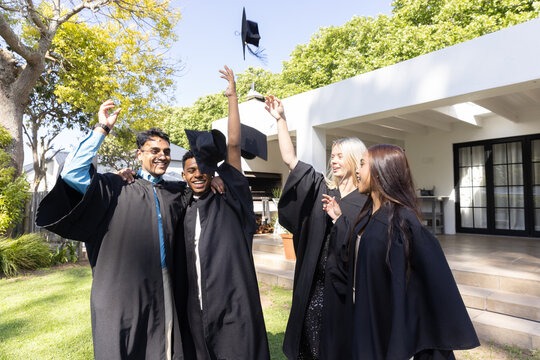 Diverse classmates in academic gowns tossing mortarboard caps celebrating graduation on lawn