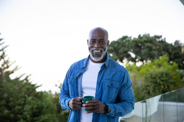 African american senior male holding green mug and smiling on balcony overlooking garden
