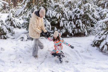 Young woman helping teenage girl stand up in snowy pine forest during playful winter walk and outdoor family leisure