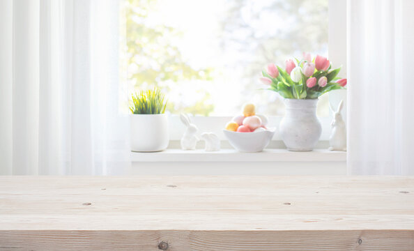 Empty wooden table on blurred background of window sill with Easter tulip flowers, decorative rabbits and colorful eggs