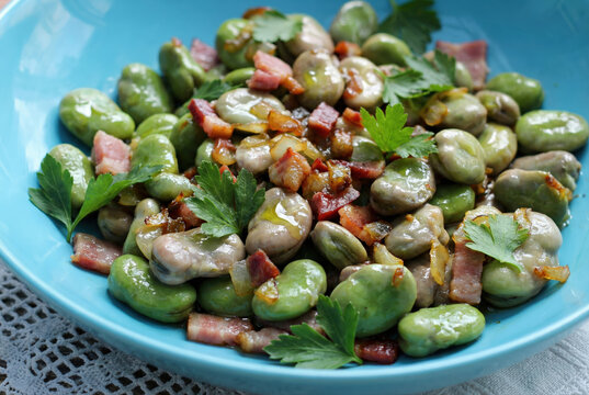 Plato de habas con jam&oacute;n y panceta crujiente decorado con perejil fresco servido en un cuenco azul sobre mantel de ganchillo