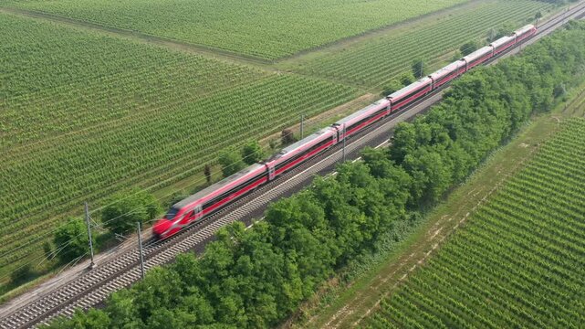 The red high-speed train swiftly glides over lush vineyard landscapes, merging modern technology with Italian countryside charm. Aerial view captures its motion.