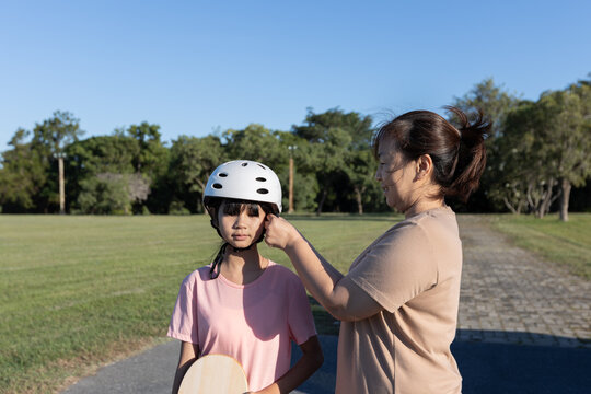 Asian mother adjusts a safety helmet for her young daughter during outdoor activity in a park, showing child safety, parental care, and family lifestyle.