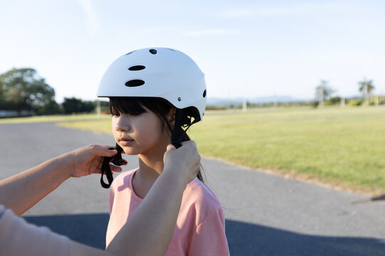 Close up of an Asian girl wearing a safety helmet while a parent adjusts the strap outdoors, showing child safety, parental care, and protection.