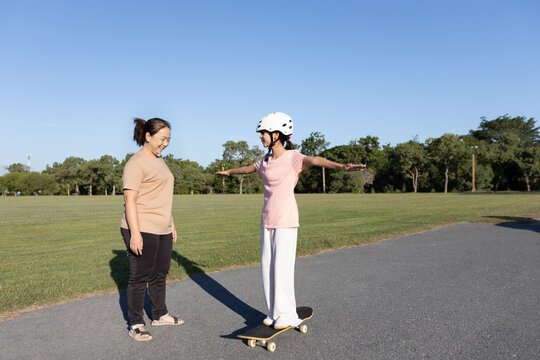Asian mother watches her daughter balancing on a skateboard outdoors while wearing a safety helmet, showing parental supervision, child safety, and learning activity.